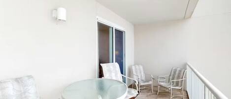 A balcony with a glass table and white chairs, a white wall, and a window with a view.