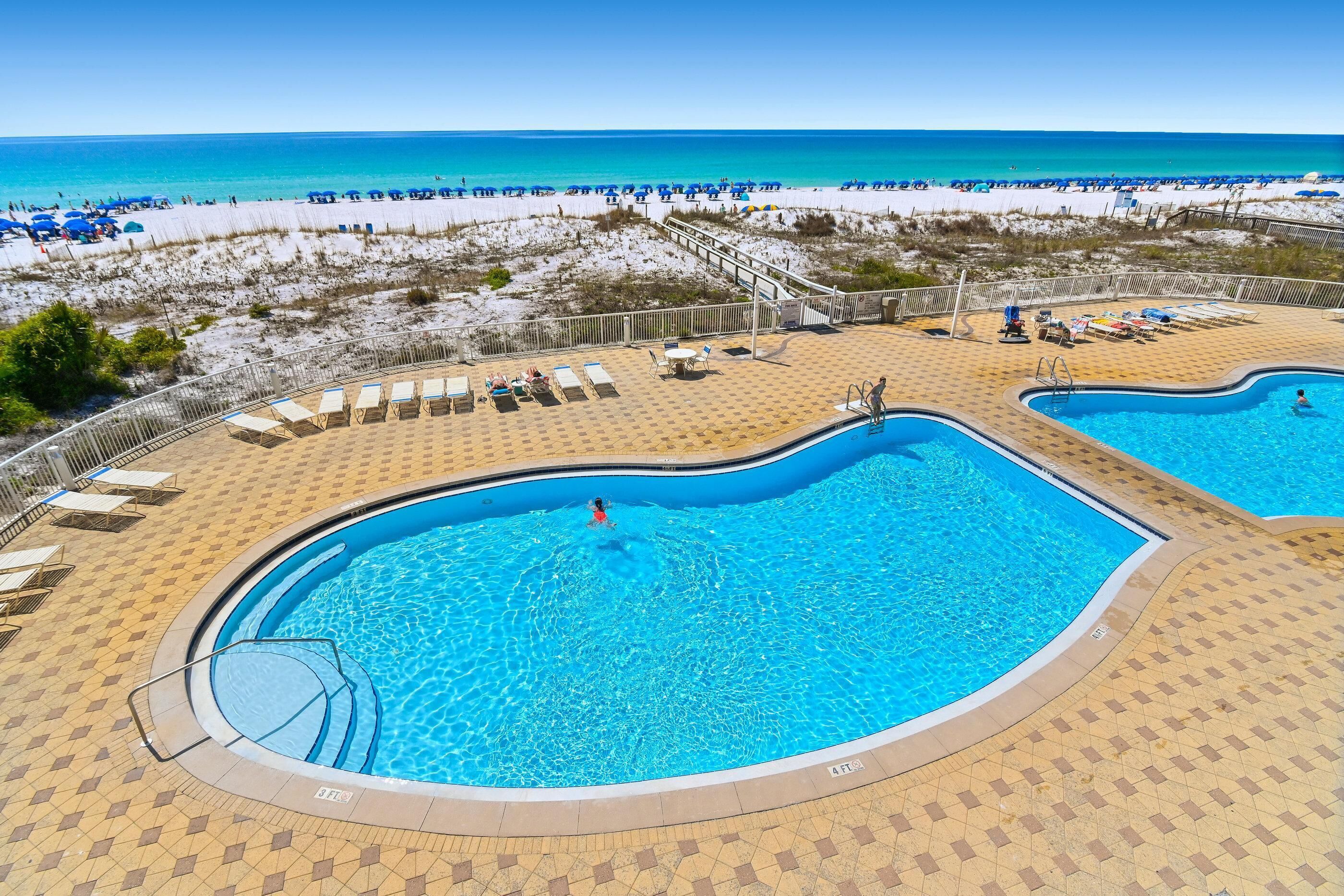 A pool area with two swimming pools, sun loungers, and a beach view.
