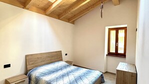 Une chambre avec un plafond en pente, des poutres en bois, un lit recouvert d’une couette bleue et blanche, une table de chevet, une commode, un meuble en bois et une fenêtre avec vue.