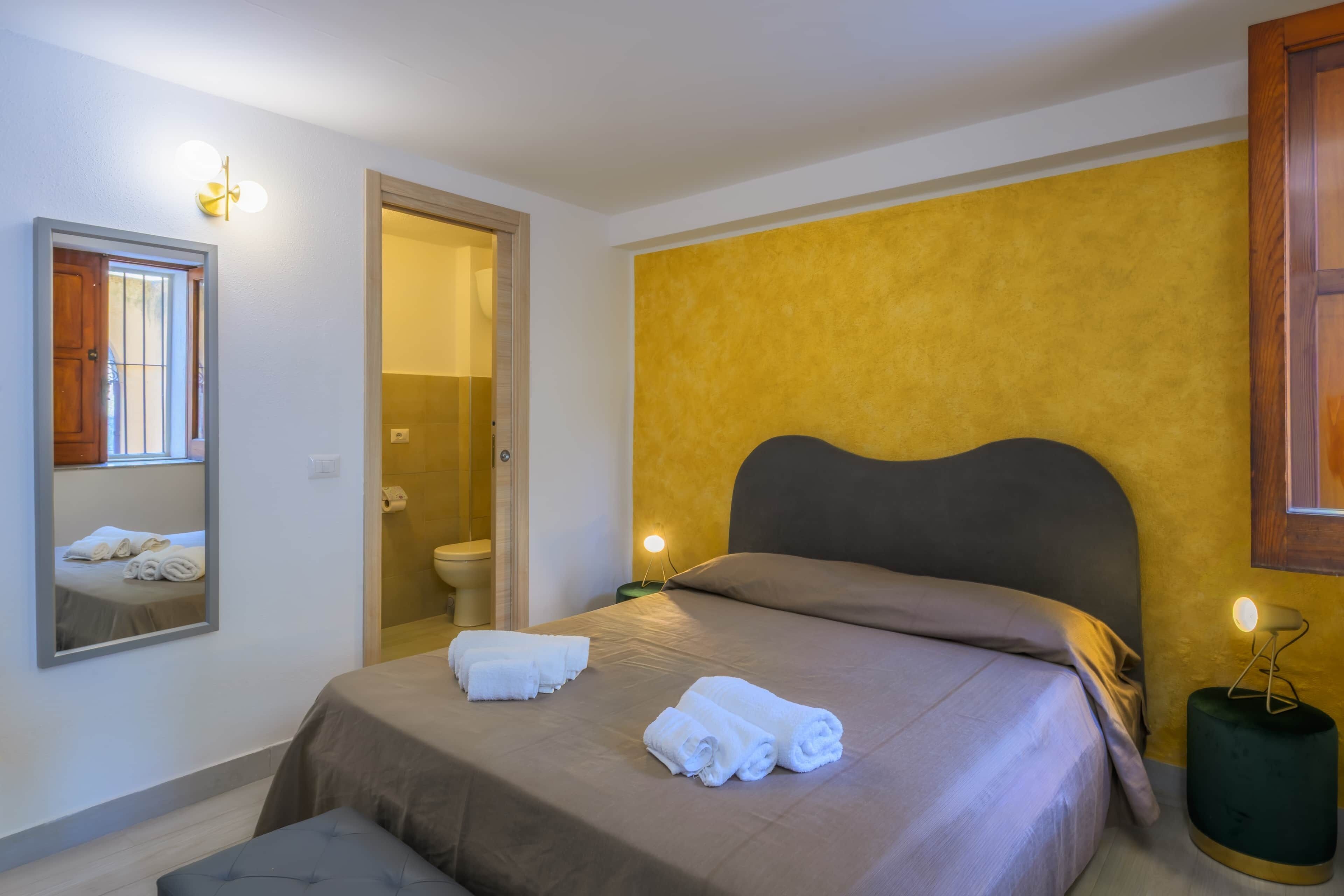 A hotel room with a bed, a mirror, a bathroom, and a yellow accent wall.