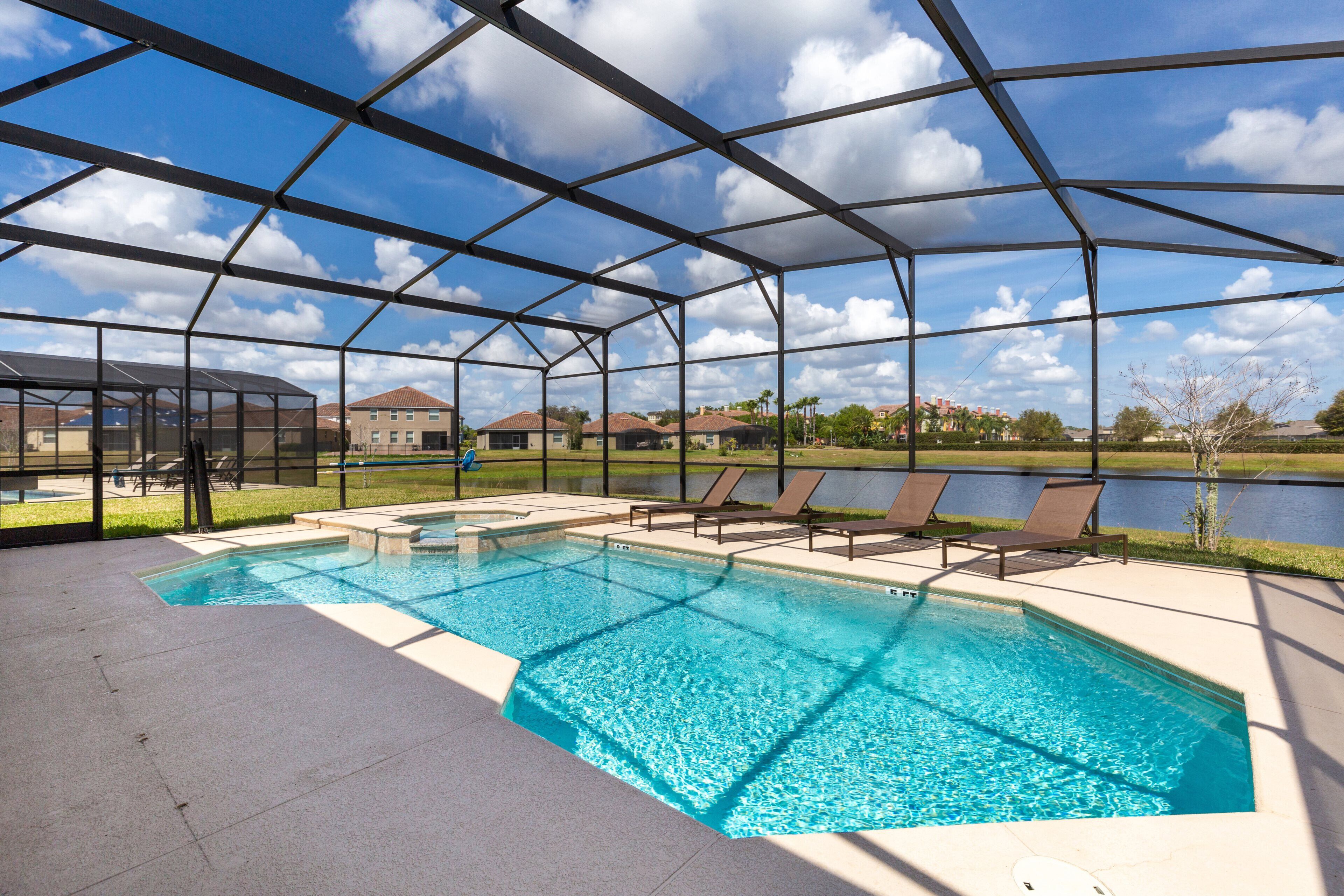 A covered outdoor swimming pool with lounge chairs and a view of a lake and buildings in the background.