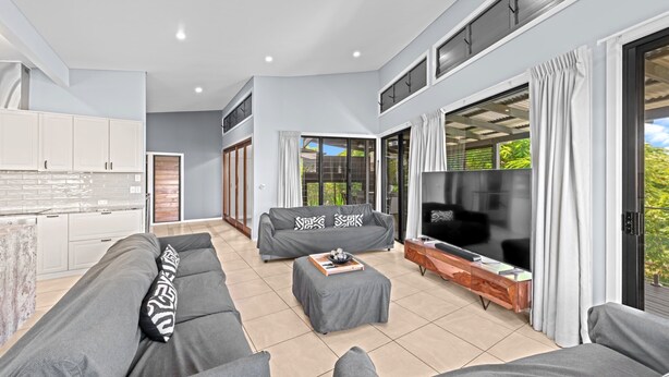 A modern living room with a grey sofa, a coffee table, and a TV. There is a kitchen area with white cabinets and a large window. - Family-sized Retreat With Private Garage (Brisbane)
