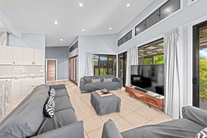 A modern living room with a grey sofa, a coffee table, and a TV. There is a kitchen area with white cabinets and a large window. - Family-sized Retreat With Private Garage (Brisbane)