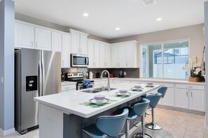 A modern kitchen with white cabinets, stainless steel appliances, a central island with bar stools, and a window with blinds. - Last Minute Disney Getaway! Luxury Villa with Private Pool. Book Now! #31556 (Orlando)