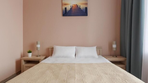 A hotel room with a large bed, two bedside tables, a nightstand with a lamp, a small plant, and a painting of a sunset over a pier on the wall. - Nika Apart Hotel  (Riga)