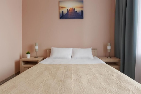 Una habitación de hotel con una cama grande, dos mesitas de noche, una mesita con una lámpara, una planta pequeña y un cuadro de una puesta de sol sobre un muelle en la pared.