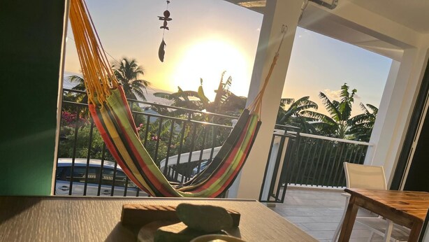 A hammock on a balcony with a view of palm trees and the sun setting in the background. - Villa EKRIN (Bouillante)