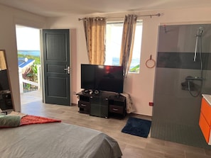 A hotel room with a bed, a TV, a shower, and a balcony with a view of the sea. - Villa EKRIN (Bouillante)