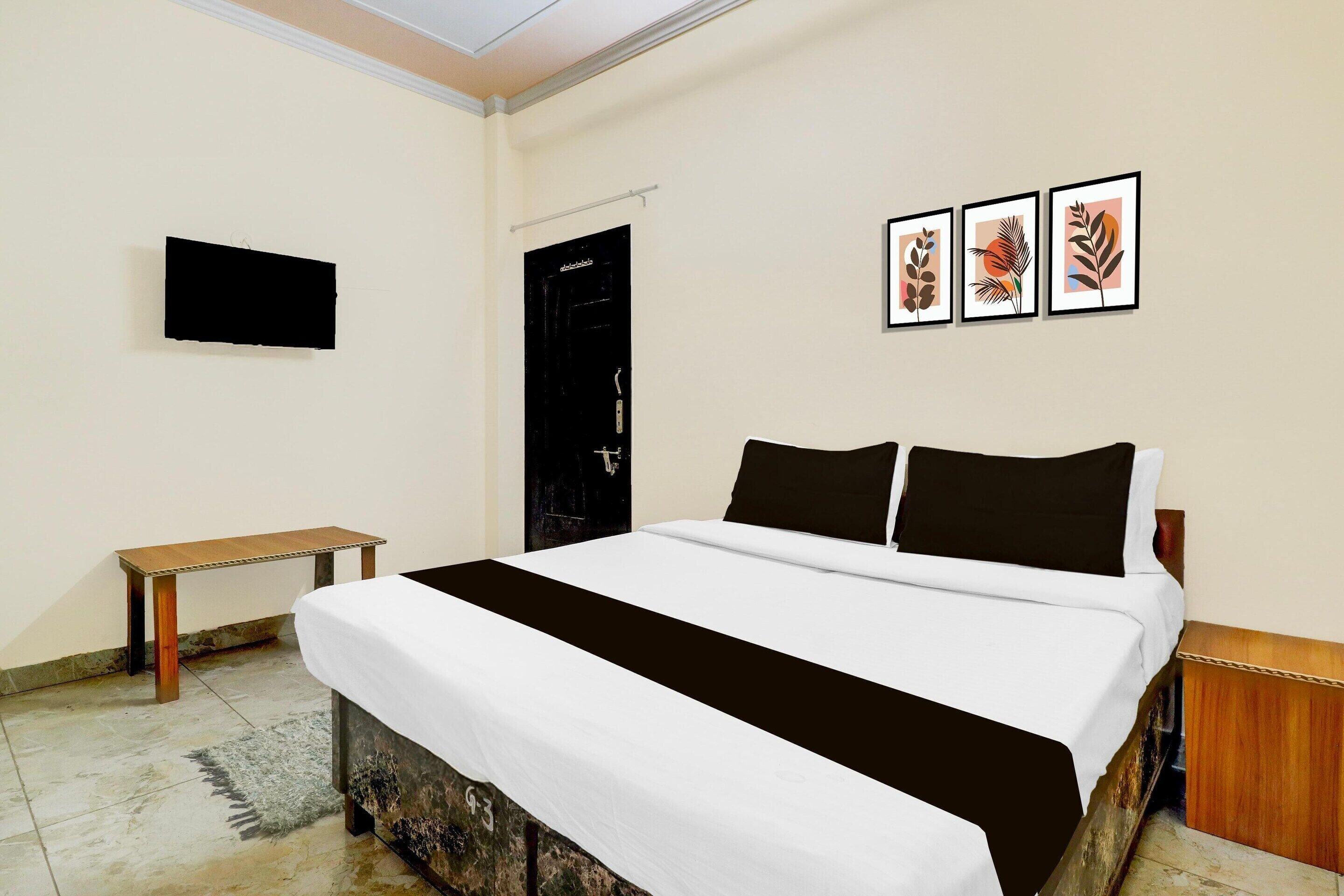 A bedroom with a bed, a bedside table, a flat-screen TV, and three framed artworks on the wall.