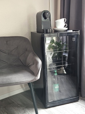 Ein kleiner Kühlschrank mit einer Wasserflasche darin, einer weißen Tasse darauf und einer kleinen Pflanze an der Tür.