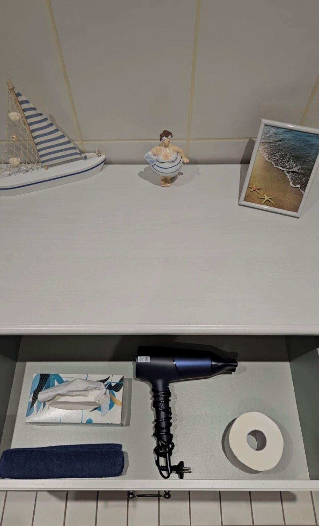 A shelf with a model ship, a figurine, a framed picture, a hairdryer, a towel, and a roll of toilet paper.