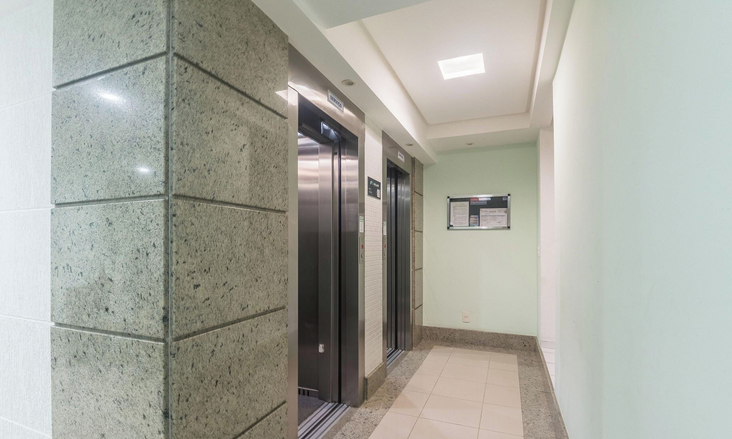 Corredor com dois elevadores, piso ladrilhado e placa fixada na parede.
