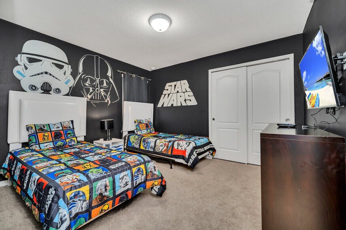 A bedroom with two beds, Star Wars themed bedding, a large wall mural, a TV, and a door.