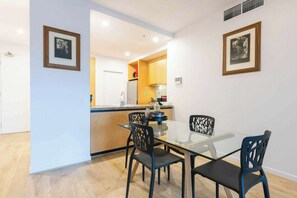 A dining area with a glass table and four chairs, a kitchen in the background, and two framed pictures on the walls. - Southbank 2BDR With Views Gym Pool And Sauna (Southbank)
