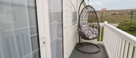 A balcony with a hanging chair, a white railing, and a view of a fenced area.