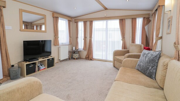 A spacious living room with a flat-screen TV, beige sofas, and large windows with curtains. - 11, Beachfields (Rhyl)
