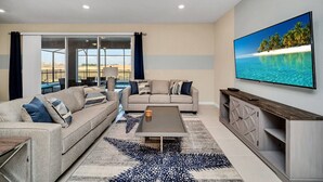 Una sala de estar con sofá, mesa de centro y televisor. Se ve un ventanal con cortinas y vistas a la playa.