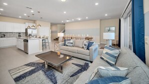 Una sala de estar moderna con sofá, mesa de centro y área de cocina con armarios blancos y un refrigerador.