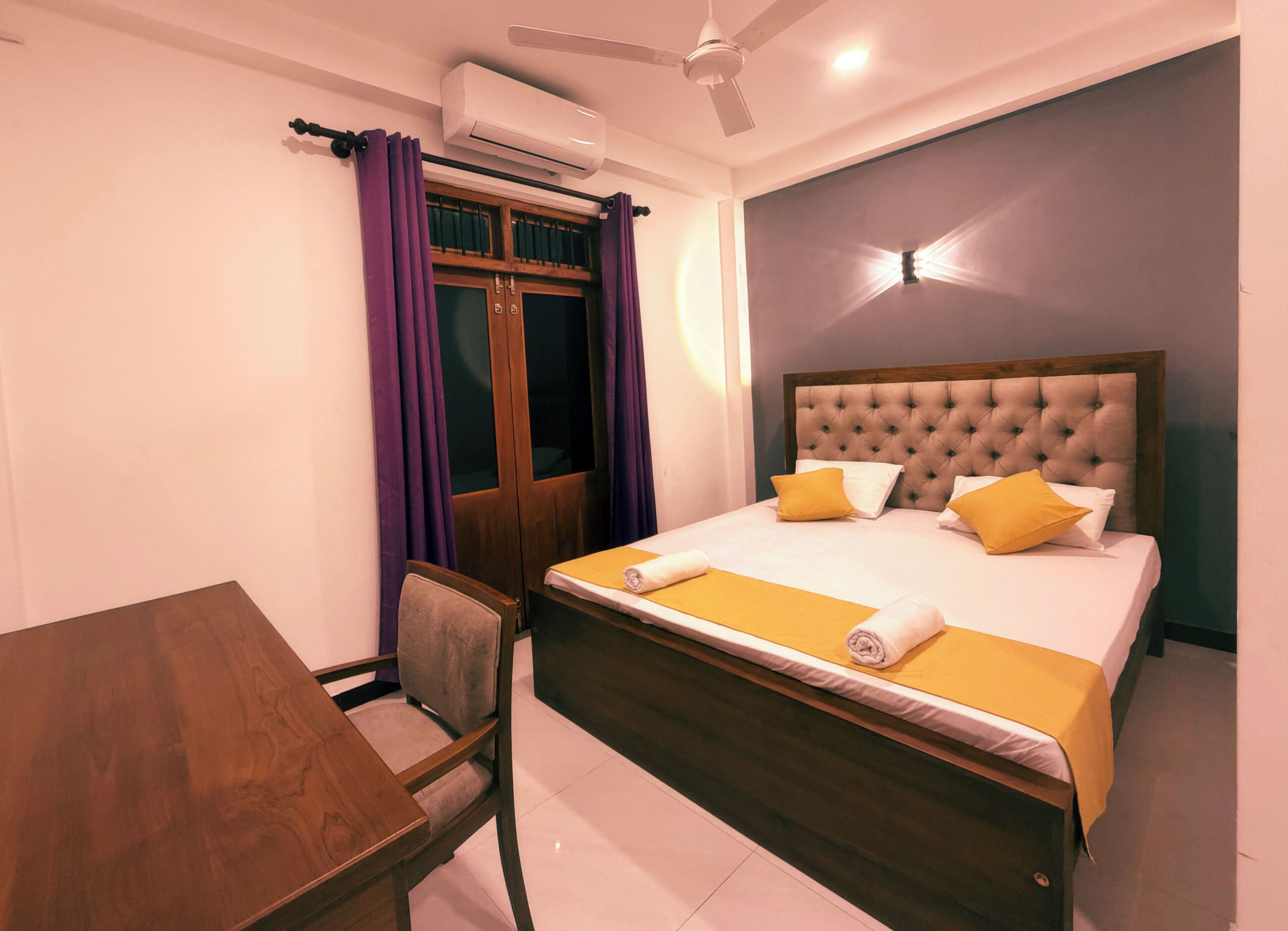 Habitación de hotel con cama, escritorio, silla, aire acondicionado y ventilador de techo.