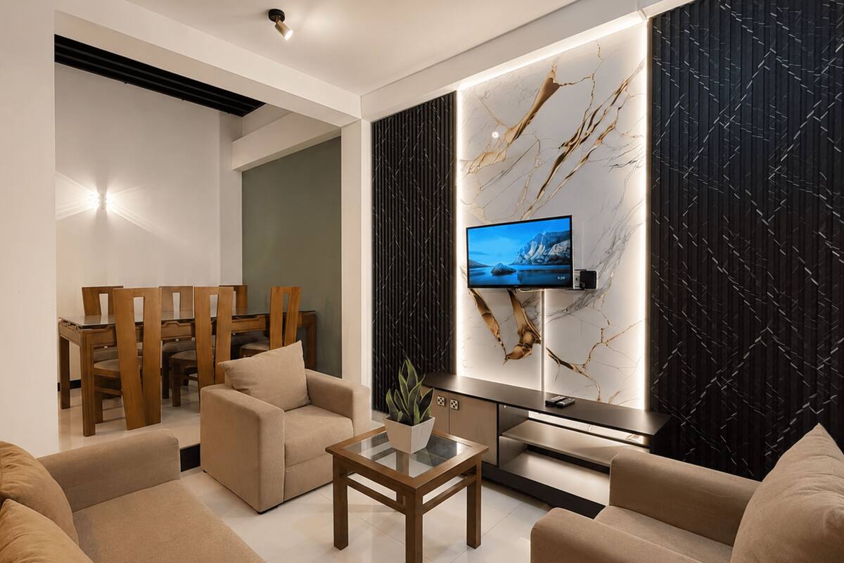 Una sala de estar moderna con un televisor de pantalla plana montado en la pared, una zona de comedor con muebles de madera y un conjunto de sofás.