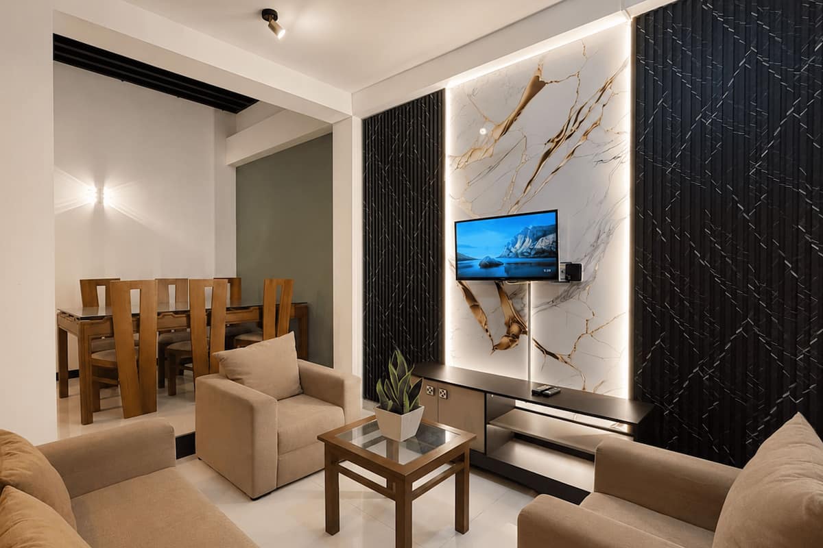 A modern living room with a flat-screen TV mounted on a wall, a dining area with wooden furniture, and a sofa set.