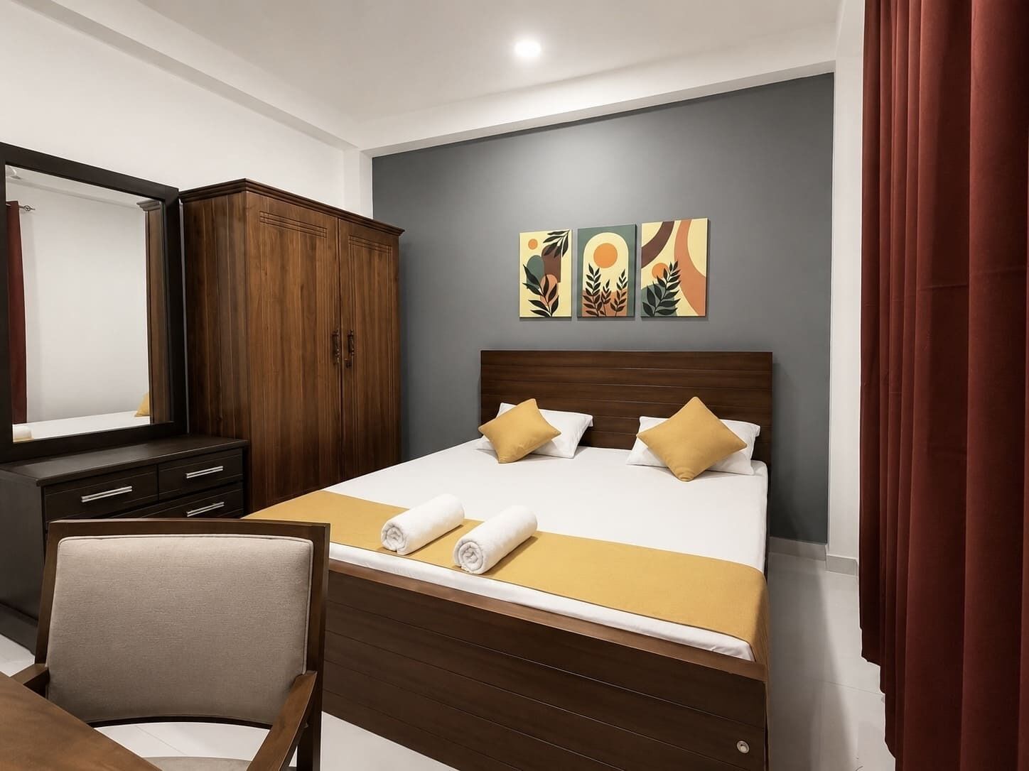 A hotel room with a bed, a wooden wardrobe, a desk, and a chair. There are three wall art pieces above the bed.