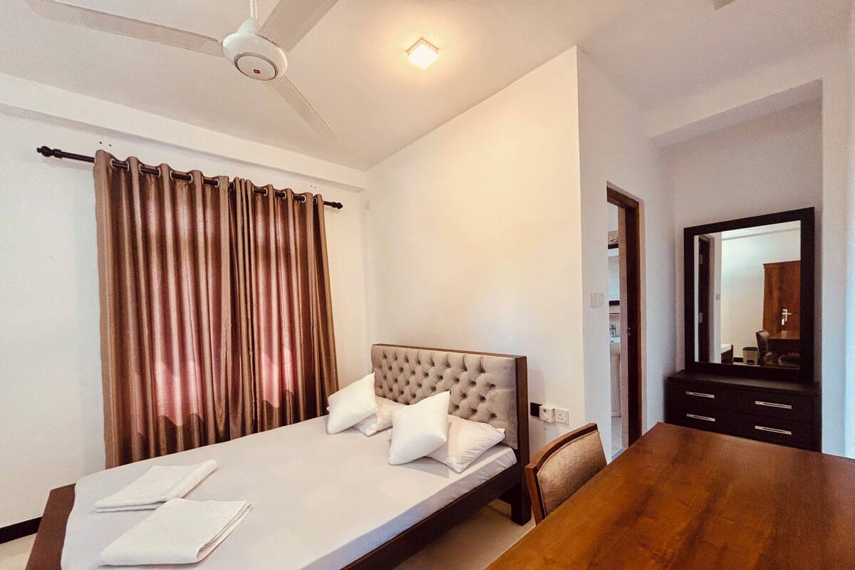 Una cama individual con cabecera, una mesa de madera, un ventilador de techo y un espejo en la pared.