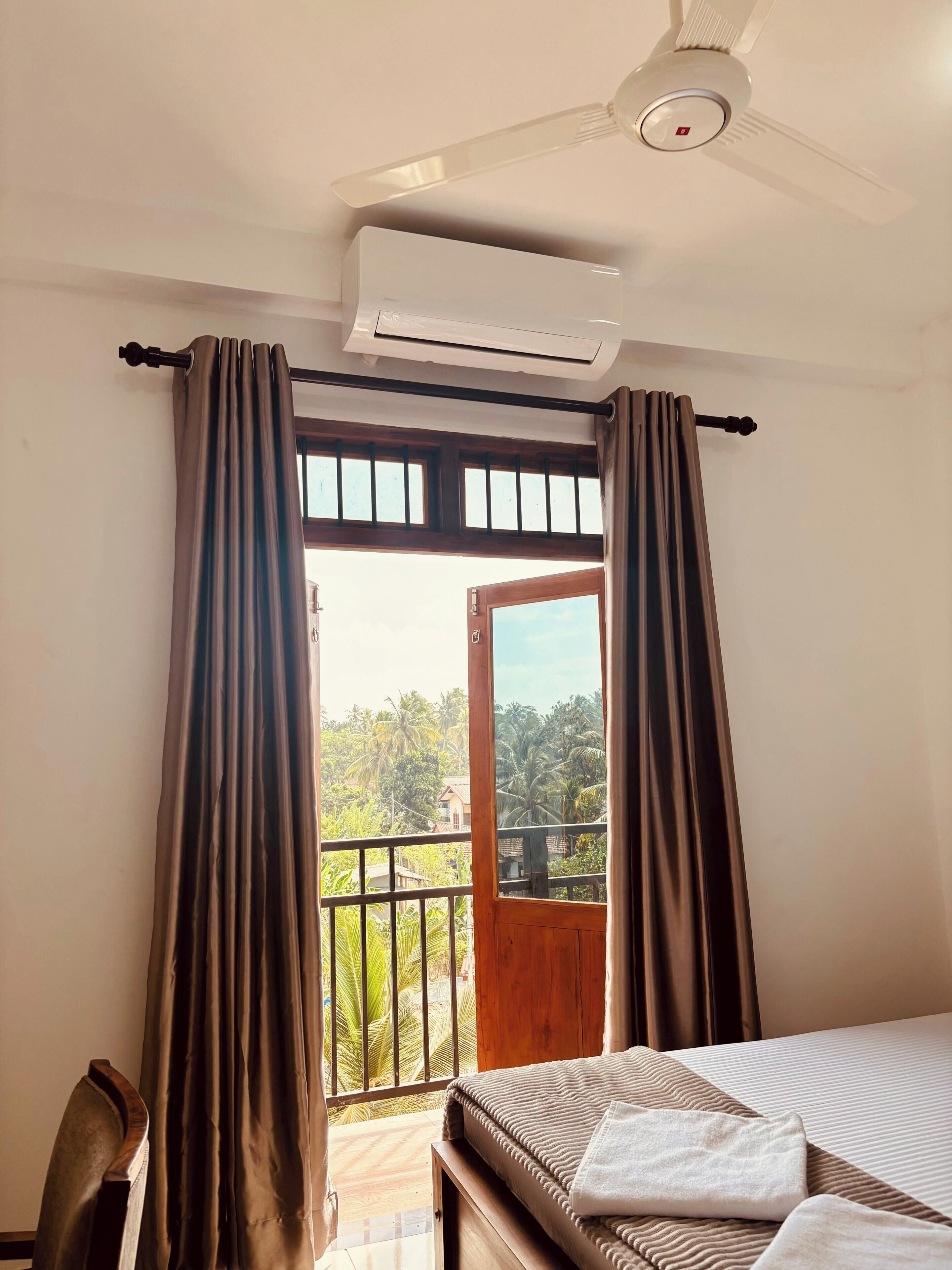 A hotel room with a bed, a ceiling fan, an air conditioning unit, and a balcony with curtains.
