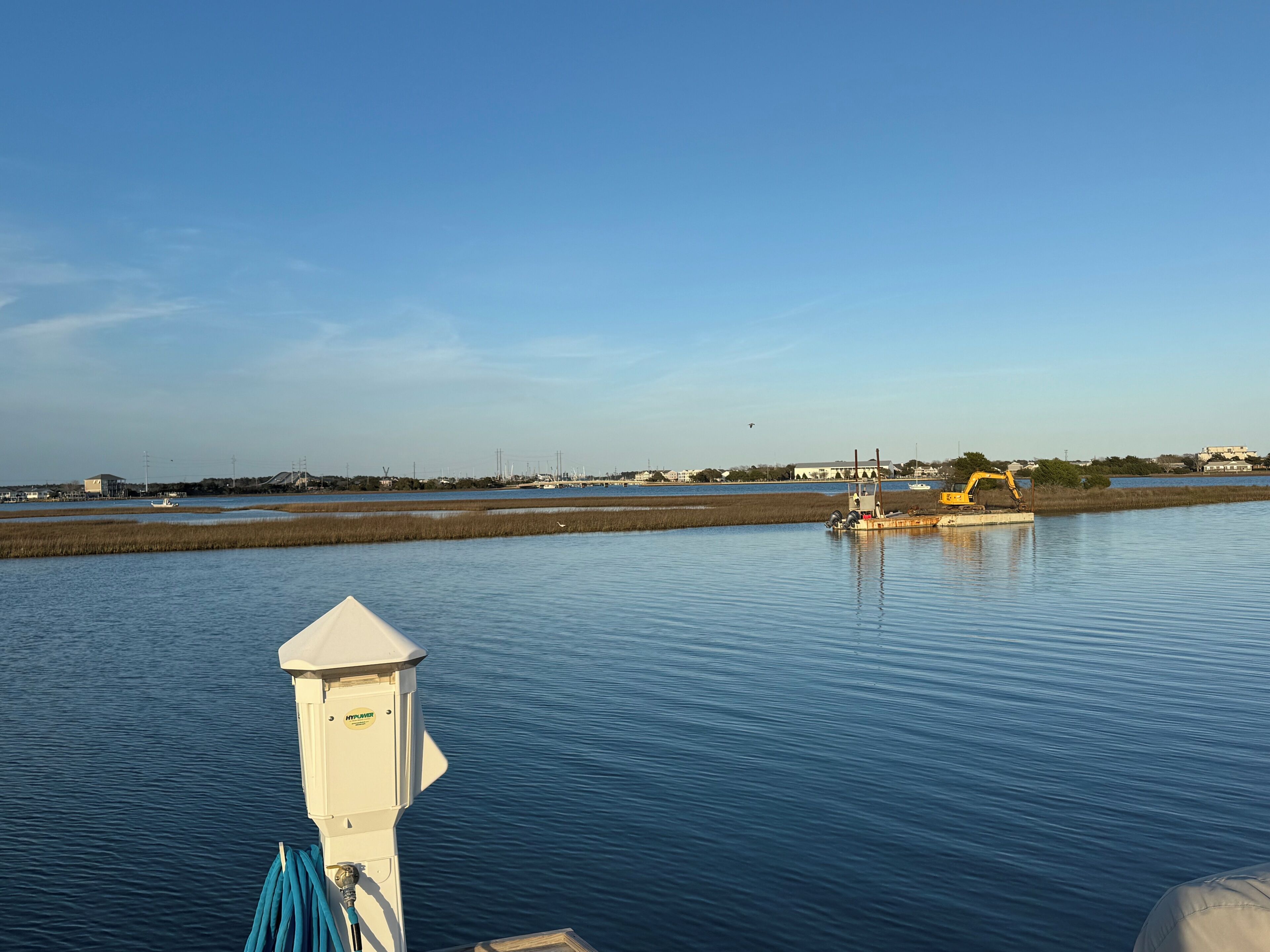Un paysage paisible au bord de l’eau, avec une structure blanche, une excavatrice jaune et un bateau en arrière-plan.