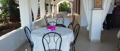 A covered outdoor dining area with white tables and chairs, a potted plant, and a framed picture on the wall.