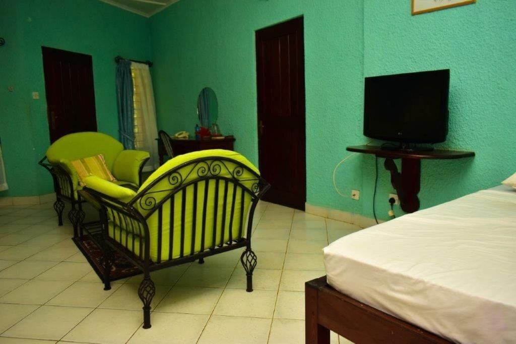 Een kamer met een bed, een groene fauteuil, een tafeltje met een televisie en een raam met gordijnen.