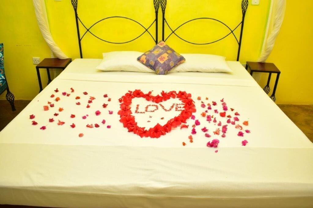 Una cama con una colcha blanca, una decoración con forma de corazón hecha de pétalos de rosa que deletrea 'LOVE', y una sola almohada.