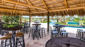 An outdoor patio area with round tables and bar stools, a pool, and a view of a landscaped garden. - Last Minute Disney! Private Pool, Hot Tub, Game Room & Themed Bedrooms. Book Now! #118573 (Orlando)