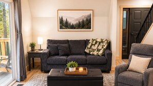 Een woonkamer met een donkergrijze bank, een bijpassend voetenbankje, een kamerkleed met patroon, een ingelijst schilderij aan de muur, en een trap die naar een deur leidt.