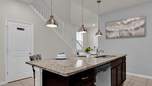 Une cuisine dotée d’un plan de travail en granit, d’un évier en acier inoxydable et d’un lave-vaisselle. On aperçoit un escalier au fond et un tableau au mur.