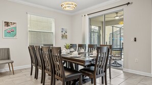 Un coin repas avec une table en bois dressée pour six personnes, entourée de chaises en bois foncé. On y trouve deux fenêtres avec des stores, un lustre et des œuvres d’art accrochées aux murs.