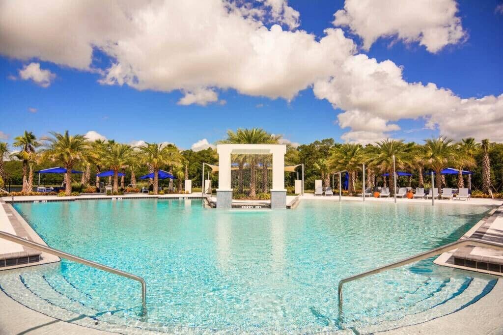 A clear blue pool with a central water feature, surrounded by palm trees and lounge chairs under blue umbrellas.