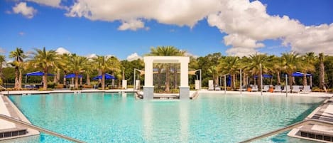 A clear blue pool with a central water feature, surrounded by palm trees and lounge chairs under blue umbrellas.