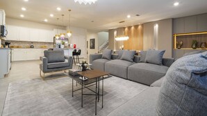 Ein modernes Wohnzimmer mit einem grauen Sofa, einem Couchtisch und einem grauen Sessel. Im Hintergrund ist eine Küche mit weißen Schränken und einem Edelstahlkühlschrank zu sehen.