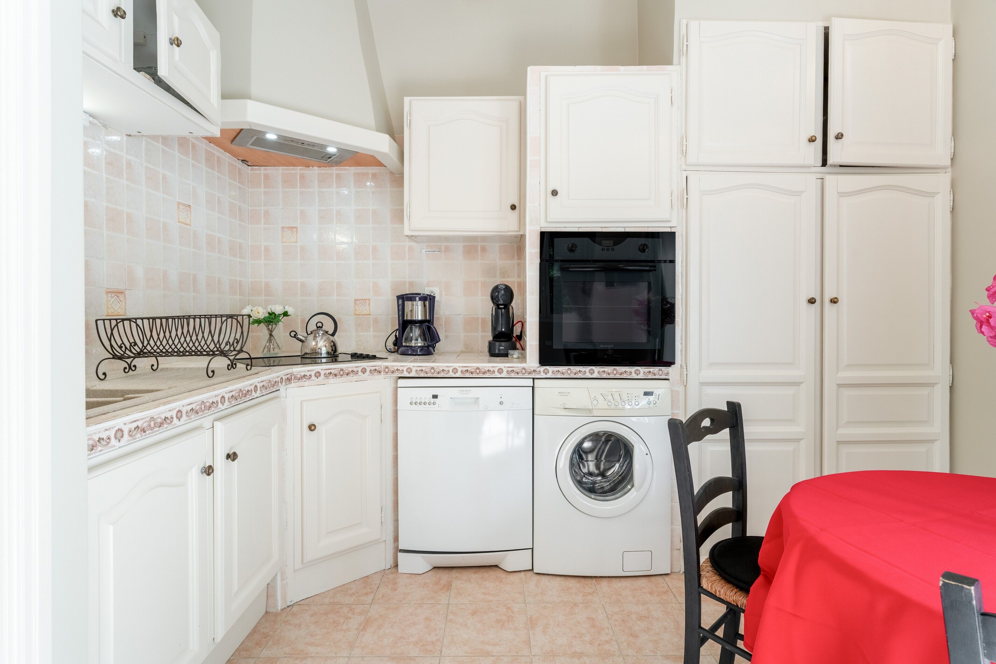 Una cucina compatta con mobili bianchi, una lavatrice, una macchina da caffè e un tavolo rosso.
