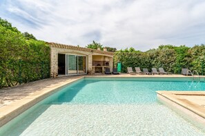 A swimming pool with a stone-tiled edge, a stone building with a glass door, and a green fence.