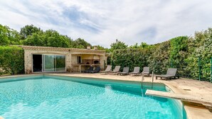 A swimming pool with lounge chairs, a small building with a fireplace, and a green fence.