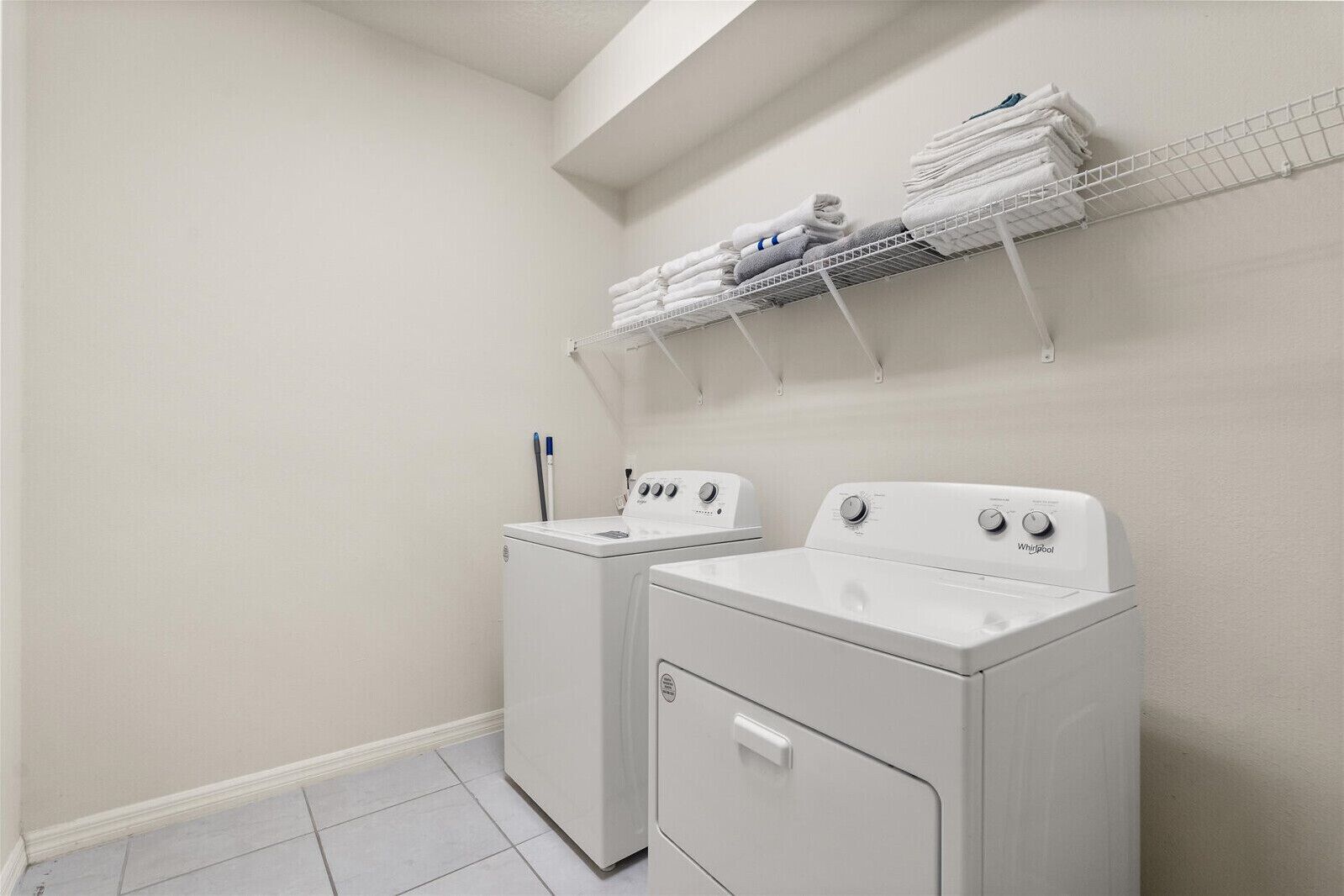 A laundry room with two white washers and dryers, a metal shelf above holding folded towels, and a white tiled floor.