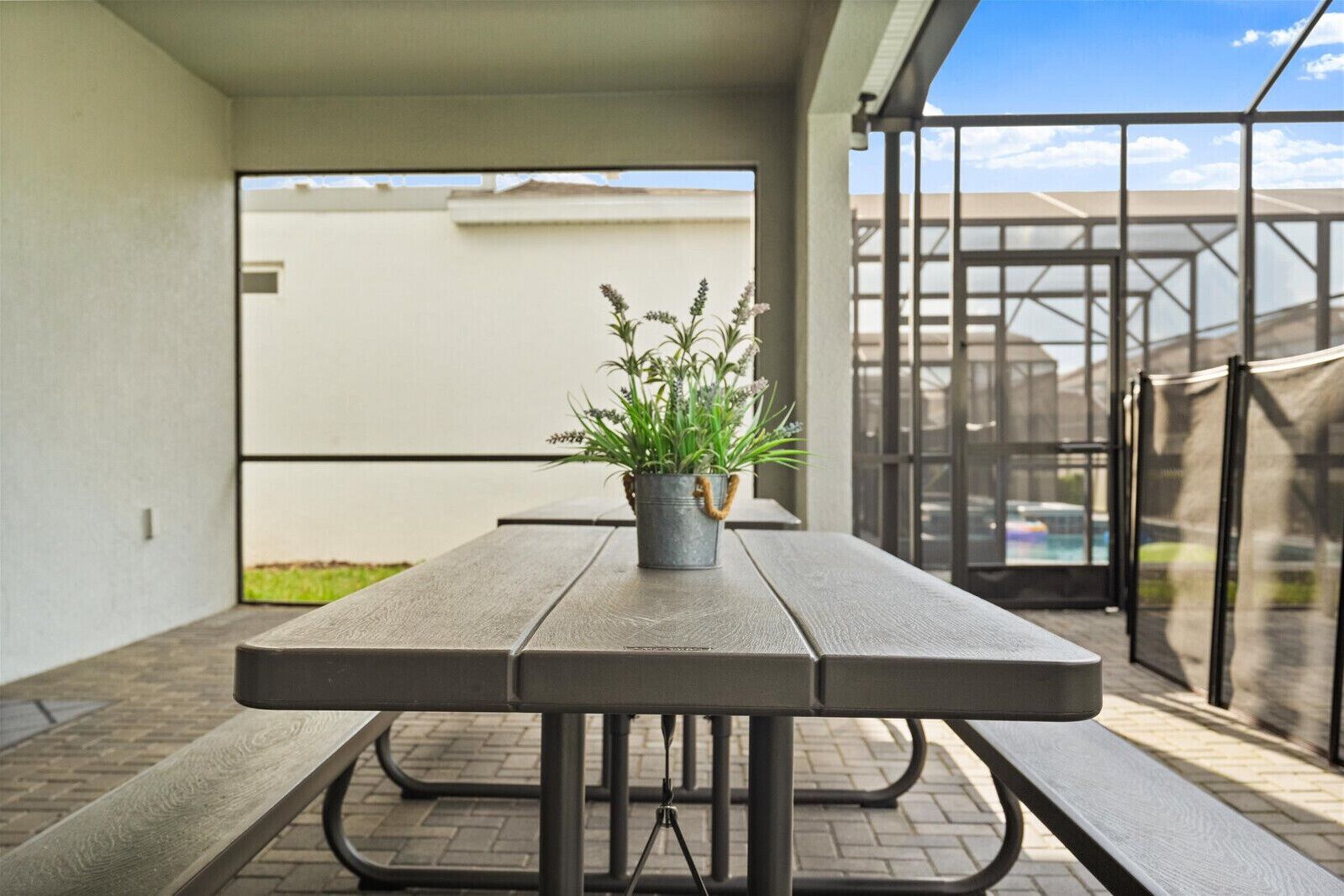 Un patio con mesa y bancos de madera, una planta en maceta y vista a una piscina a través de un ventanal.