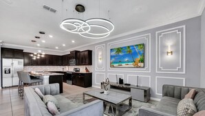 A modern living room with a grey sofa, a coffee table, and a TV wall-mounted above a kitchen area with dark cabinets and a central island. - Last Minute Disney! Private Pool, Hot Tub, Game Room & Themed Bedrooms. Book Now! #WIR437 (Orlando)