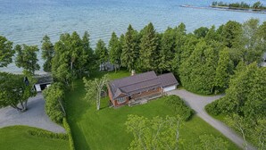 A large house with a garage, surrounded by a well-maintained lawn and trees, near a body of water. - Private Lake Simcoe Retreat | Sandy Beach, Sunsets, Kayaks & Ice Fishing (Beaverton)