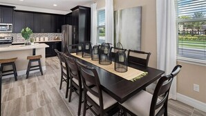A modern kitchen with dark cabinetry, a central island with stools, and a dining area with a long table and chairs. - Last Minute Disney Getaway! Townhouse with Private Pool. Book Now! #136377 (Orlando)