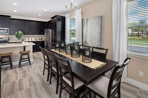 A modern kitchen with dark cabinetry, a central island with stools, and a dining area with a long table and chairs. - Last Minute Disney Getaway! Townhouse with Private Pool. Book Now! #136377 (Orlando)