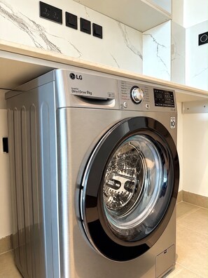 Una lavadora de carga frontal moderna LG Inverter DirectDrive de 9kg, con panel de control digital, ubicada en una cocina con encimeras de mármol y salpicadero de azulejos.