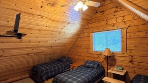 A cozy cabin with wooden walls and a sloped ceiling. There are two beds with plaid bedding, a small table, and a lamp. A ceiling fan is visible, and a window with blinds is on the wall.